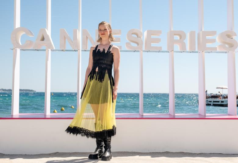  Kuvassa on Roosa Söderholm seisomassa puvussa, jossa on musta hihaton yläosa ja alaosana läpinäkyvä keltainen tyllipuolihame, jonka alla on lyhyt puolihame. Taustalla on valkoisella jalustalla valkoisia pystylautoja ja teksti Canseries. Taustalla on sinistä merta. 