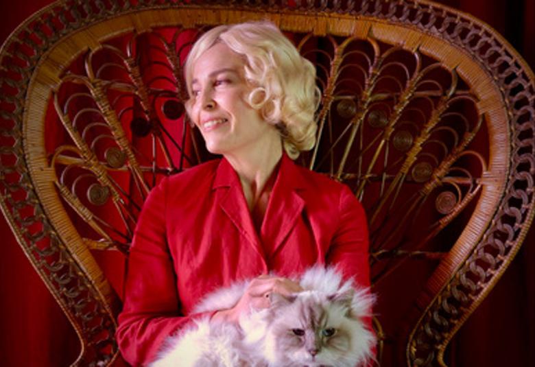 Kuvassa on Irina Björklund istumassa isossa korituolissa ja hänellä on valkoinen kissa sylissään. Björklundilla on vaaleat hiukset ja punainen mekko päällään. Takaseinä on punainen.