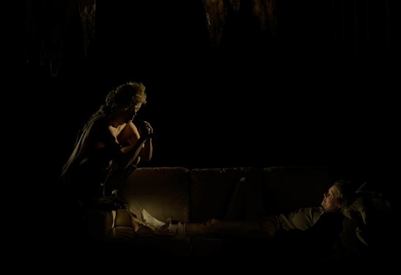 Kuvassa on tummalla pohjalla juuri ja juuri näkyvissä vasemmalla naishahmo kumarassa ja lattialla makaamassa mies.