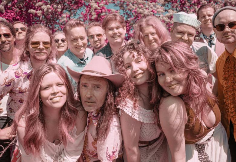 Kuvassa on puolivartalokuvassa runsaasti nuoria esiintyjiä tiiviisti vierekkäin ryhmänä koko kuva-alalla. Kuvan sävy on vaalean punainen.