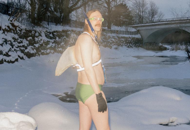 Kuvassa on Stig menossa lumisessa maisemassa sillan ali vitaavaan veteen uimaan. Hänella on valkoiset siivet selässään ja vaalean vihreät uimahousut sekä mustat hansikkaat.