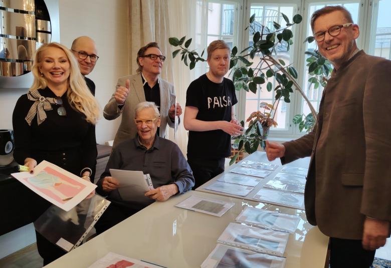 Kuvassa ovat suuren pöydän ääressä seisomassa on vasemmalla viisi henkilöä ja edessä oikealla seisoo yksi mies. Pöydällä on taideteosten luonnoksia A4 koossa.