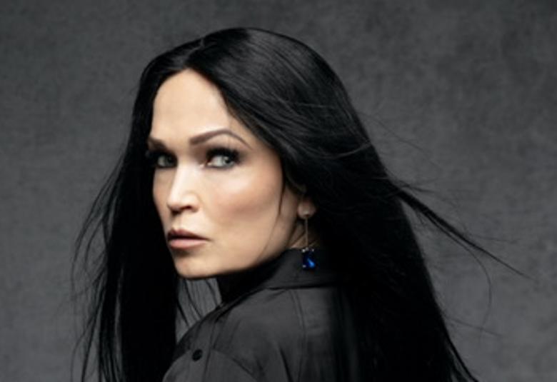 Kuvassa on Tarja Turunen puolivartalokuvassa ja hän katsoo olkapään yli. Hänellä on pitkät, tummat hiukset ja musta jakku. Tausta on tumman harmaa.