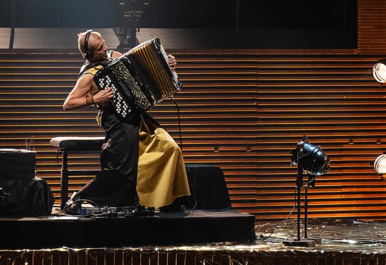 Kuvassa on vasemmalla jalustalla sivuttain Kimmo Pohjonen keltaisissa leveissä housuissa soittamassa harmonikkaa. Takana on ruskeaa vaakasuoraa rimaa.