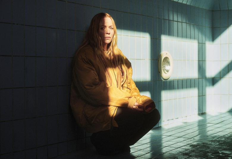   Kuvassa on Paula Vesala kyykussä ja vasen puoli kuvasta on tummaa ja oikealle tulee auringon valoa. Vesala näkyy puolittain. Taustalla on isojen lasi-ikkunoiden nurkka.