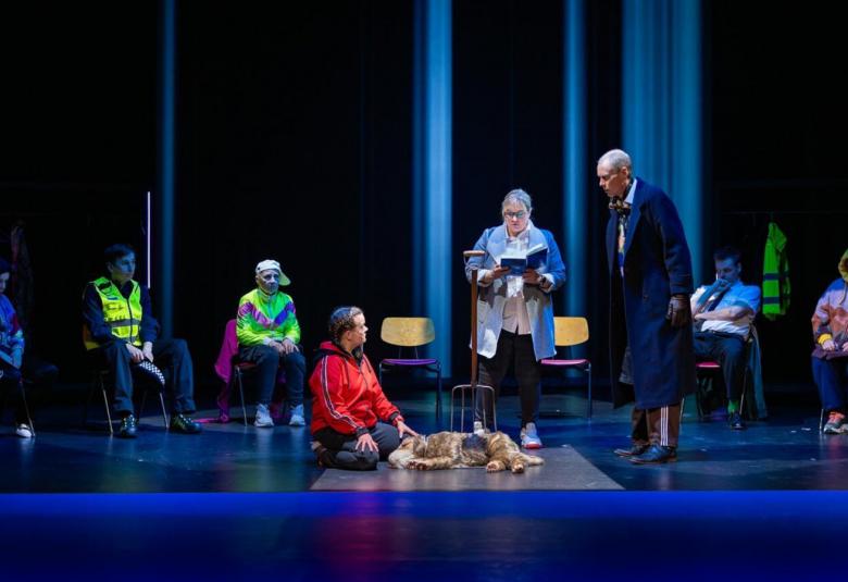 Kuvassa on tummalla näyttämöllä edessä mies punaisessa pusakassa polvillaan ja hänen edessään on vaalean ruskea koira. Hänen vieressään seisoo nainen lukemassa kirjasta tekstiä ja hänellä on vaalean sininen jakku. Hänen vierellään oikealla seisoo mies tummassa takissa. Vasemmalla istuu kolme henkilöä ja oikealla kaksi pusakoissa.