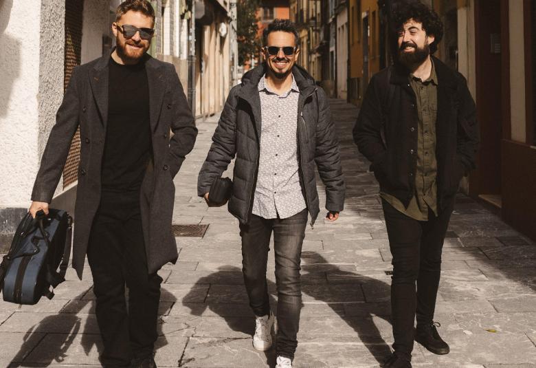 Kuvassa on kolme nuorta miestä kävelemässä kapeaa talojen reunustamaa katua. Heillä n mustat vaatteet paitsi keskimmäisellä harmaa paita.