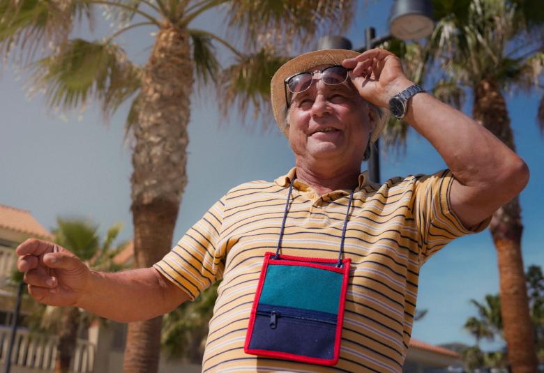 Kuvassa on Tom Lindholm kuvattuna yläviistoon. Hänellä on karttalaukku kaulassaan ja hän pitää aurinkolaseista kiinni vasemmalla kädellään. Taustalla on palmuja, vaaleita rakennuksia ja sinistä taivasta.