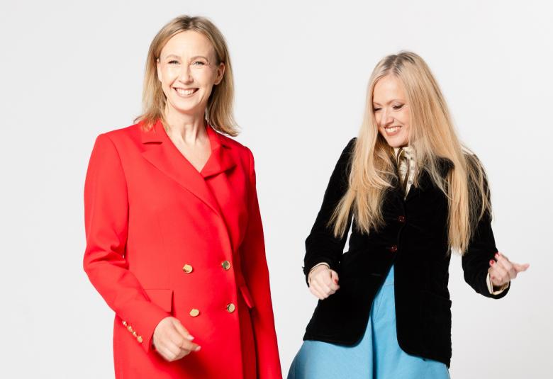 Kuvassa on vasemmalla punaisessa takkipuvussa Heli Meklin ja oikealla on Eveliina Talvitie jammailemassa mustassa jakussa ja vaalean sinisessä mekossa. Tausta on vaalea.