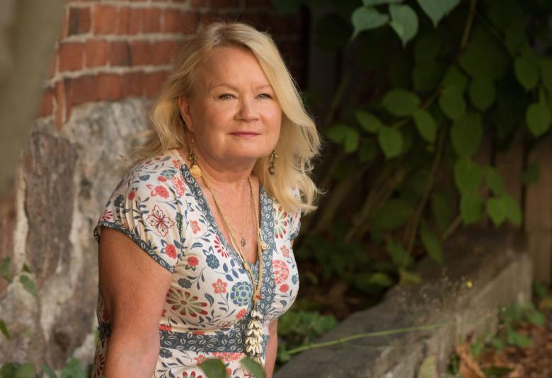 Kuvassa on vasemmalla Kaarina Griffiths istumassa puolivartalokuvassa vaalean kukallisessa mekossa. Taustalla on tiiliseinää ja vihreää pensasta.
