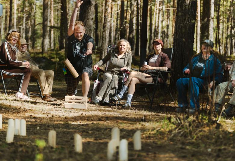 Kuvassa on metsässä istumassa nurmikon takana ja siinä näkyy miehiä ja naisia vasemmalla olevan karavaanerin nokka. Edessä on mölkkypalikoita pystyssä. 