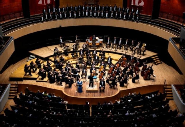 Kuvassa on Sinfonia Lahti ylhäältä kuvattuna soikealla puisella esiintymislavalla soittamassa. Ylempänä näkyy parvekkeella yleisöä ja edessä permannolla. Kuva on osa kirjan kannesta.