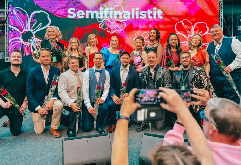 Kuvassa ovat kahdessa rivissä Tangomarkkinoiden semifinalistit ja heistä ensimmäisessä rivissä olevat nojaavat toiseen polveensa. Taustalla on punaisia kukkia Tangomarkkinoiden isossa lakanassa. Edessä on yleisöä ottamassa valokuvia.