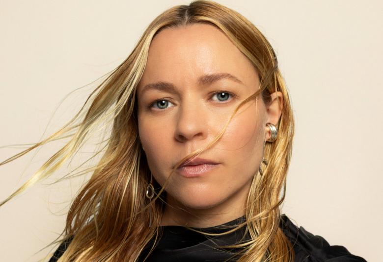  Kuvassa on Paula Vesala kasvokuvassa. Hänellä on pitkät, vaaleat hiukset ja ne liehuvat tuulessa. Tausta on vaalea.