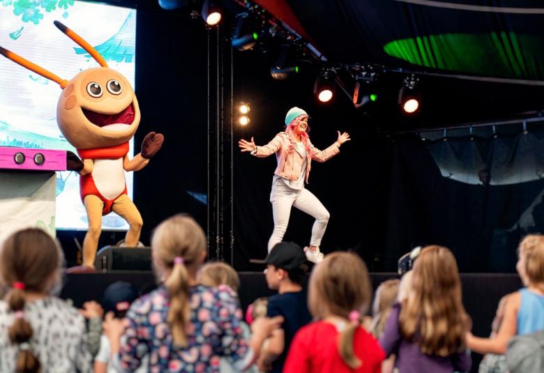 Kuvassa on esiintymislavalla vasemmalla Kikattava Kakkiainen, joka on oranssinen otus isoine päineen ja tuntosarvineen. Silla on punainen paita päällään. Lavalla oikealla on naislaulaja, jolla vaaleat vaatteet ja hän tekee loikkaa kädet levitettyinä.