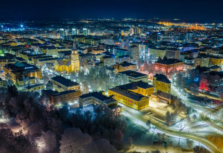 Kuvassa on sinertävän pimeää Oulun keskustaa ylhäältä päin kuvattuna ja kuvassa on kaupunkia valaistuna.