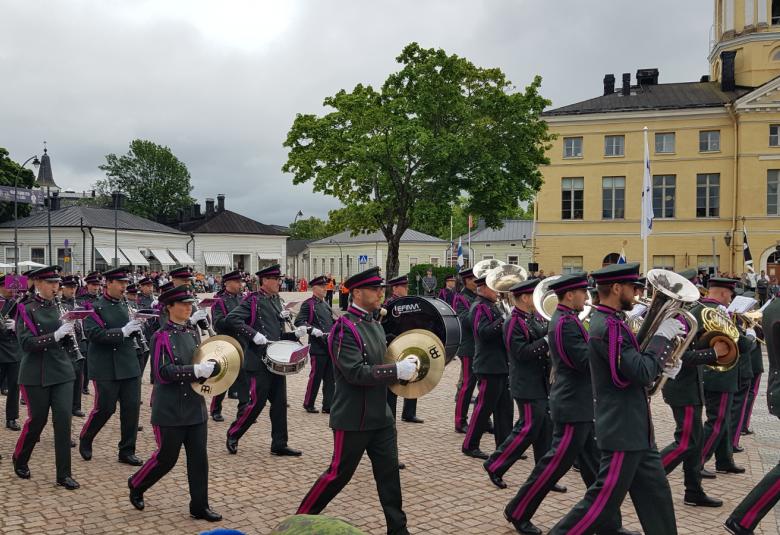 Kuvassa on tumma pukuisia sotilassoittajia riveissä marssimassa ja soittamassa. Taustalla keskellä näkyy keltainen Haminan raatihuone.