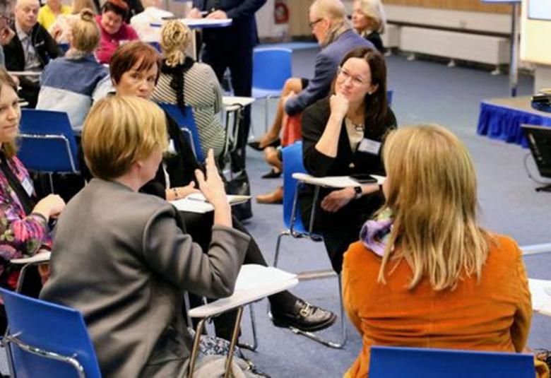 Kuvassa on naisia ryhmissä ja pöydät käännettyinä siten että voivat keskustella keskenään. Tuolit ovat sinisiä ja lattia harmaa.