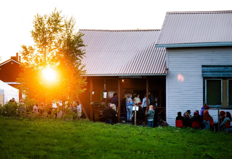 Kuvassa on valkoinen talo, jonka päässä on latomainen ja sen takaa paistaa ilta-aurinko. Rakennuksen edessä on ihmisiä ja vihreää nurmikkoa. 
