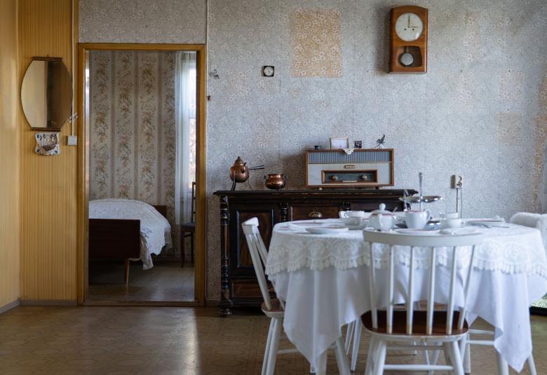Kuvassa on huone, jossa on etualalla pöytäkalusto, jossa on valkoiset tuolit ja valkoinen pöydän peittävä liina. Taustalla on harmaatapettista seinää ja seinällä on seinäkello.