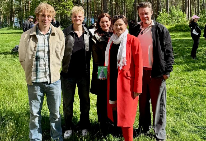 Kuvassa ovat seisomassa vasemmalta kaksi miestä vaaleassa pusakassa ja farkuissa ja etualalla nainen punaisessa takissa, taustalla on nainen tummassa takissa ja oikealla on mies tummassa takissa ja punertavassa paidassa. Taustalla on nurmikkoa ja koivikko.