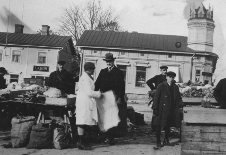 Kuvassa on etualalla torin myyntipöytä ja pöydän takana mies seisomassa. Pöydän vieressä seisoo nainen valkoisessa takissa ja mies mustassa hatussa ja lierihatussa. Oikealla on kaksi miestä ja välissä iso puulaatikko sekä sen vieressä mies. Taustalla on kaksi vaaleata, vanhaa puutaloa. Kuva on musta-valkoinen.
