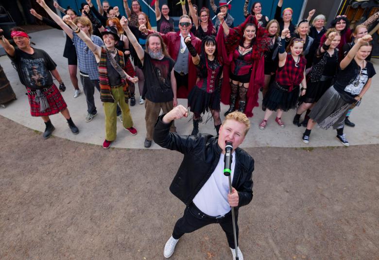 Kuvassa on ylhäältä kuvattuna nuori mies laulamassa mikin edessä ja hänellä musta takki ja housut sekä valkoinen t-paita. Takana on suori joukko laulajia ja näyttelijöitä käsi pystyssä laulamassa.