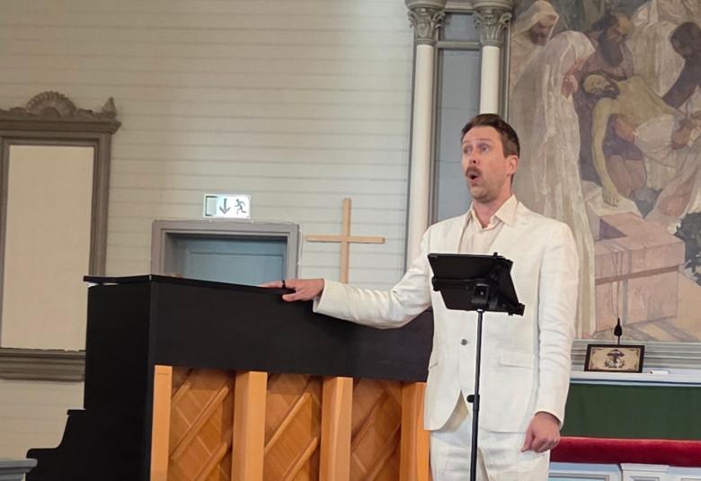 Kuvassa on Waltteri Torikka laulamassa oikealla valkoisessa puvussa ja hän pitää oikealla kättään pianon päällä. Pianosta näkyy ruskea, puinen takaosa