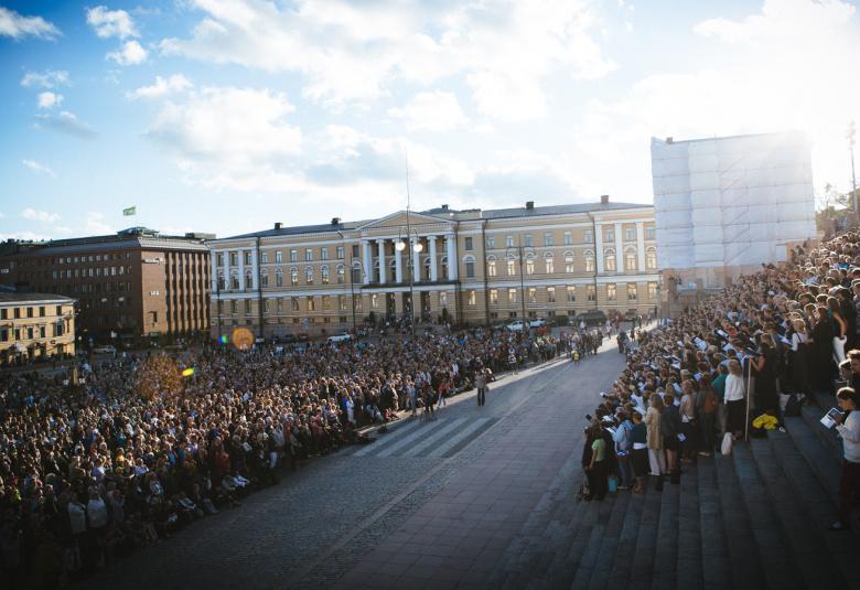 Kuvassa on Senaatintorilla vasemmalla runsaasti kuorolaisia seisomassa ja laulamassa. Oikealla on Helsingin tuomiokirkon portailla on myös runsaasti laulajia seisomassa. Keskellä on näkyvissä tilaa ja katukivetystä.