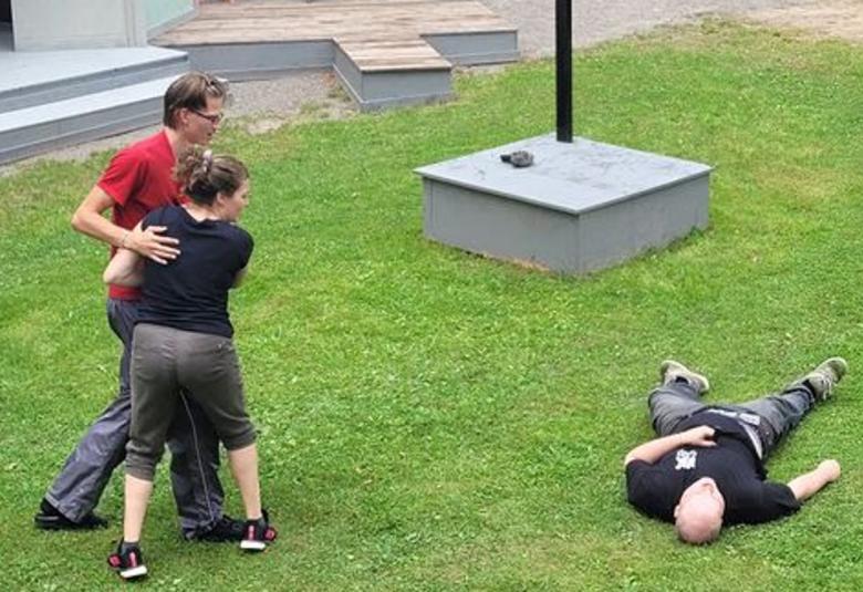 Kuvassa ovat vasemmalla nuori nainen ja mies ja he ovat jonkinlaisessa kamppailuotteessa ja oikealla katsovat maassa ruohikolla makaavaa nuorta miestä. Taustalla näkyy harmaita lavoja.