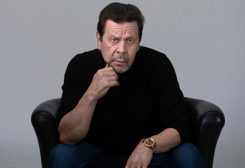 Kuvassa on Pate Mustajärvi mustassa paidassa istumassa mustassa isossa nojatuolissa kumartuneena eteenpäin. Hän nojaa oikealla kädellään leukaansa. Tausta on harmaa.