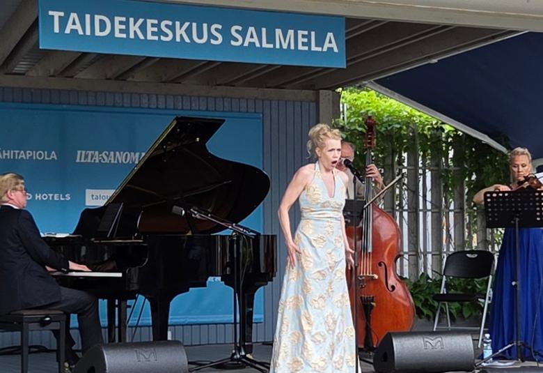 Kuvasswa on Kuvassa on vasemmalla Janne Hovi soittamassa flyygeliä ja keskellä Helena Juntunen vaaleassa, pitkässä puvussa laulamassa. Takana soittaa kontrabassoa Hannu Rantanen takana ja oikealla Maija Linkola viulua pitkässä, sinisessä puvussa. Päällä lukee Taidekeskus Salmela.