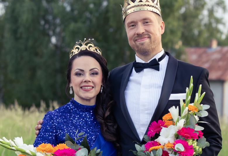 Kuvassa on vasemmalla Raija Mäntyniemi sinisessä mekossa ja vieressä oikealla Juho Punkeri mustassa takissa ja valkoisessa paidassa. Molemmilla on kukkakimput kädessään. Taustalla on metsää ja valkoinen talo.