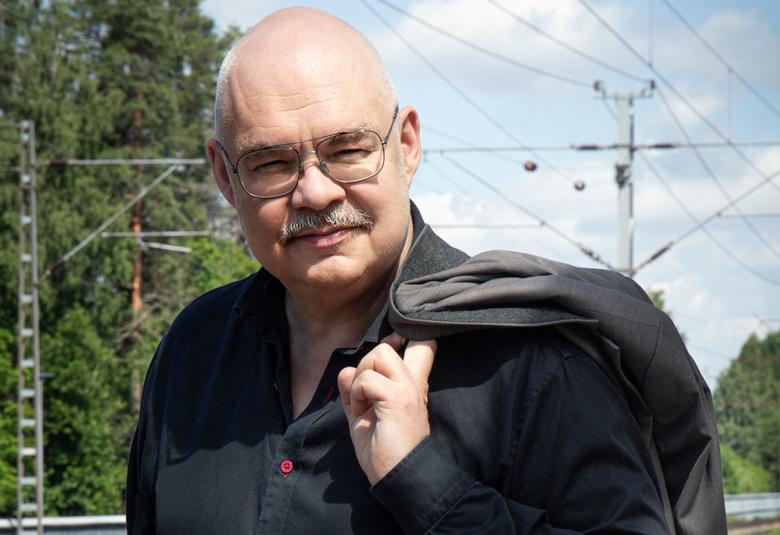 Kuvassa on Mikko Roiha puolivartalokuvassa ja hänellä on musta paita ja musta takki vasemmalla olkapäällään.  Taustalla näkyy metssä ja rautatien sähköjohtoja.