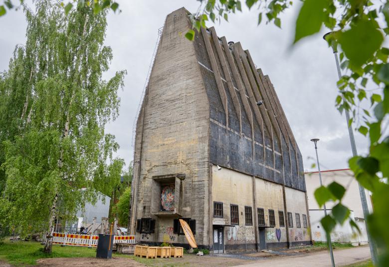 Kuvassa on keskellä harmaa, ylöspäin menevä harmaa siilo. Siinä on kattojuonteita tummalla katolla. Alhaalla on rivi ikkunoita. Molemmilla puolilla on vihreälehtisiä lehtipuita.