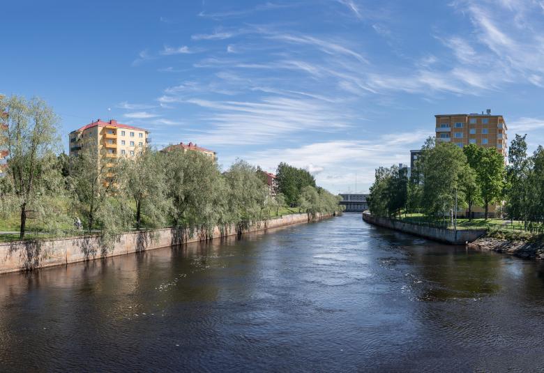 Kuvassa on keskellä joki ja sen molemmin puolin kerrostaloja, joiden edessä on lehtipuita. Takana on sinistä taivasta.