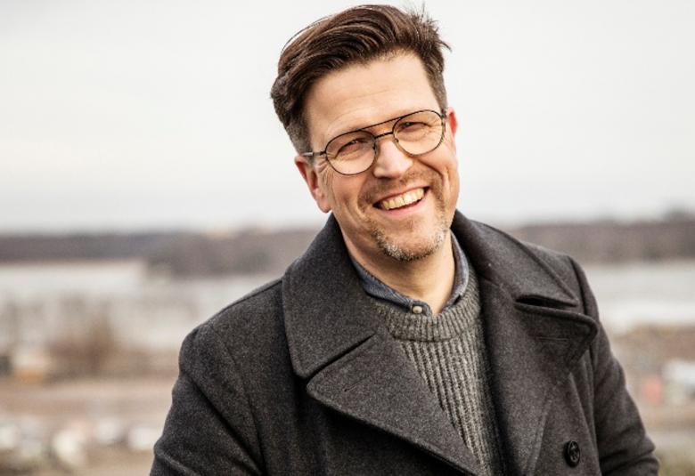 Kuvassa on Klaus Härö puolivartalokuvassa ja hänellä on ruskea takki ja sen alta näkyy harmaan ruskeata villapaitaa. Taustalla on epätarkkaa syysmaisemaa. 