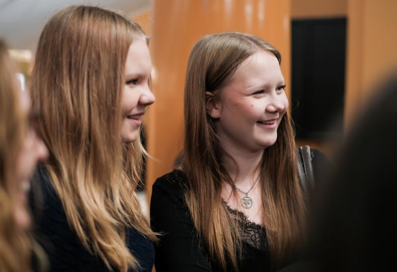 Kuvassa on kolme tyttöä puolivartalokuvassa. Heillä on kaikilla pitkät hiukset ja he seisovat vinottain. Heillä on mustat t-paidat. Taustalla on vaalean ruskeaa seinää.