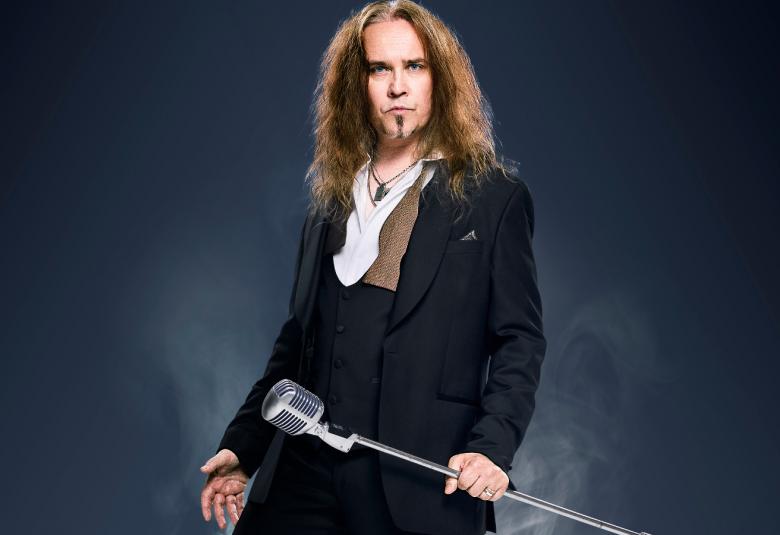 Kuvassa on keskellä on Jarkko Ahola seisomassa ja hänellä musta takki sekä valkoiset kaulukset. Hänellä on jalallinen mikrofoni oikeassa kädessään. Tausta on tumma.