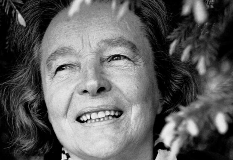 Kuvassa on Eeva Kilpi kasvokuvassa ja hänen ympärillään on kuusenoksia ja jouluvaloketjua. Kuva on musta-valkoinen.