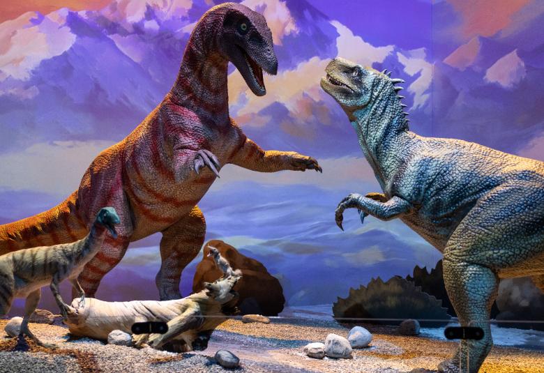 Kuvassa on kuvassa on vasemmalla violetti iso dinosaurus ja sitä vastapäätä on turkoosi iso dinosaurus. Punertavan dinosauruksen vieressä on pieni turkoosi dinosaurus. Maassa on kelopuu ja sen päässä on orava . Taustalla on sinertävän värikästä vuoristoa.