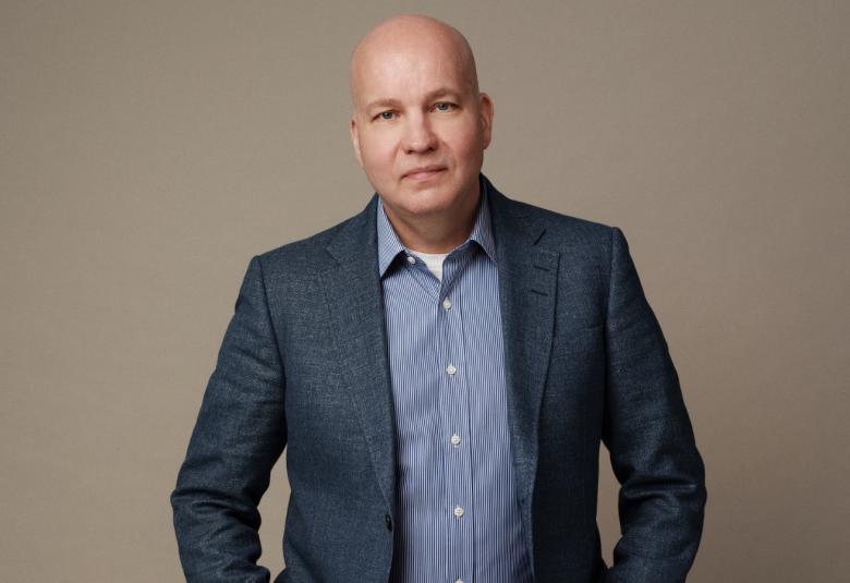 Kuvassa on Kim Väisänen seisomassa ja hänellä on sininen takki ja vaalean sininen paita. Kuvassa on harmaan ruskea tausta.