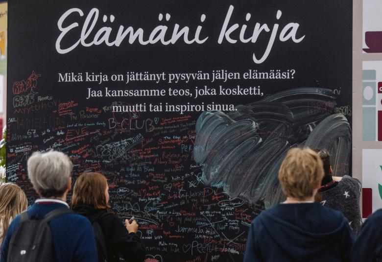Kuvassa on seinämä, jossa lukee kirjoituskirjaimin Elämäni kirja ja sen alla pientä tekstiä mustalla pohjalla. Edessä on ihmisiä katselemassa seinää.
