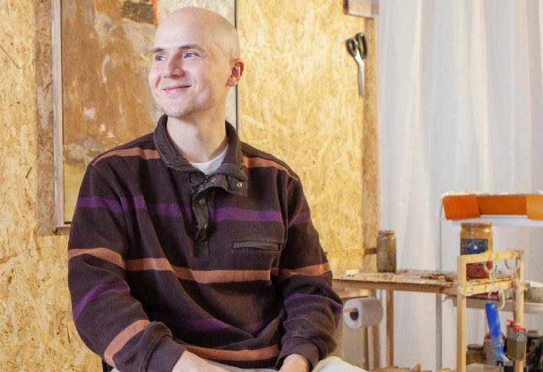 Kuvassa on Kristo Saarikoski ja hänellä on ruskearaitainen pusero päällään. Taustalla on vaaleaa seinää taideteos sekä oikealla vaaleita huonekaluja.  