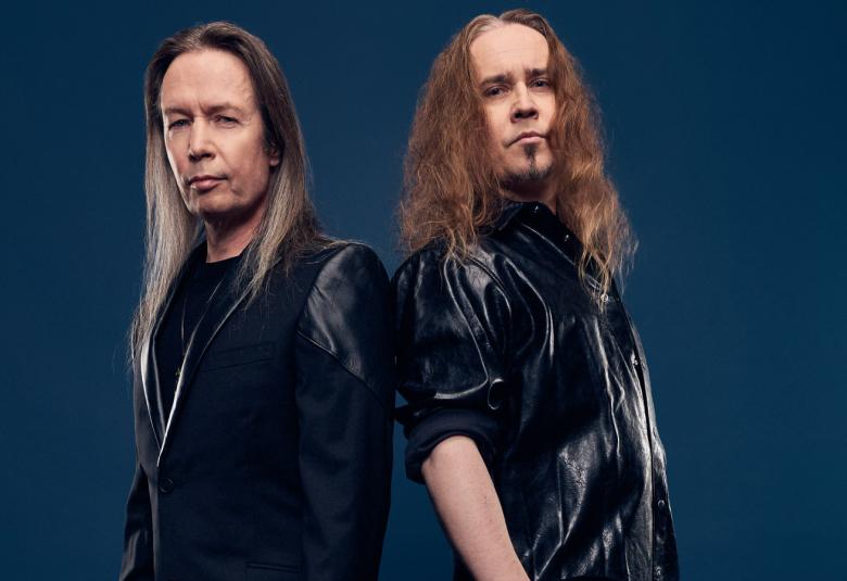 Kuvassa on vasemmalla Timo Kotipelto ja oikealla Jarkko Ahola. He ovat osittain selin ja molemmilla on mustat puserot ja pitkät hiukset.