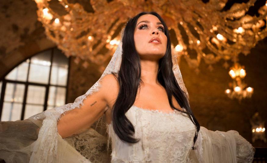 Kuvassa on keskellä Abreu valkoisessa hihattomassa pitsimekossa ja pitsihuntu päässään tummissa, pitkissä hiuksissaan sekä pitää huntua käsivarsillaan. Taustalla on ylhäällä kultainen suuri kattokruunu.  