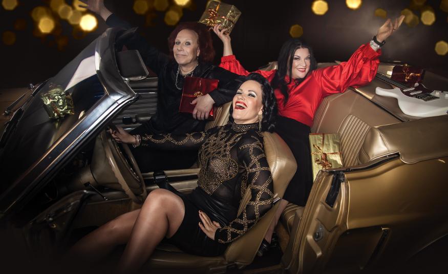 Kuvassa on etualalla Erja Lyytinen istumassa avoauton ruskealla istuimella ja hänen vieressään Kärtsy Hatakka. Takapenkillä istuu Hanna Pakarinen punaisessa puserossa molemmat kädet levitettynä ylöspäin. 