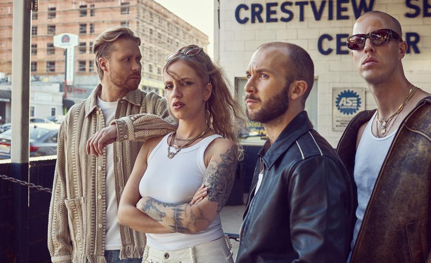 Kuvassa on Haloo Helsingin jäsenet seisomassa tilassa, jossa lukee seinässä Crestview Center. Vasemmalla on mies vaalean ruskeassa villatakissa vieressäänElli valkoisessa hihattomassa t-paidassa. Oikealla on kaksi miestä ja heillä on tummat takit päällään. Lasi-ikkunan takaa näkyy vaalean ruskea iso kerrostalo.