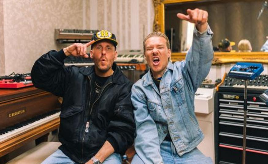 Kuvassa ovat JVG jäsenet ja vasemmalla Ville Gallella on musta nahkatakki ja Jarella vaalean sininen farkkupuku sekä vasen käsi ylhäällä. Taustalla on on soittimia ja äänitysstudio.