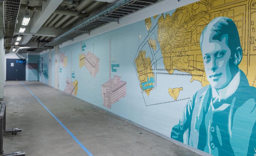 Kuvassa on käytävä, jossa on oikean puoleinen seinä ja sen pohja on vaalean sininen.  Seinällä on karttapohjamaisia kuvioita ja oikealla keltainen niemi.  Lähinnä oikealla on iso miehen rintakuva vaalean sinisessä takissa.